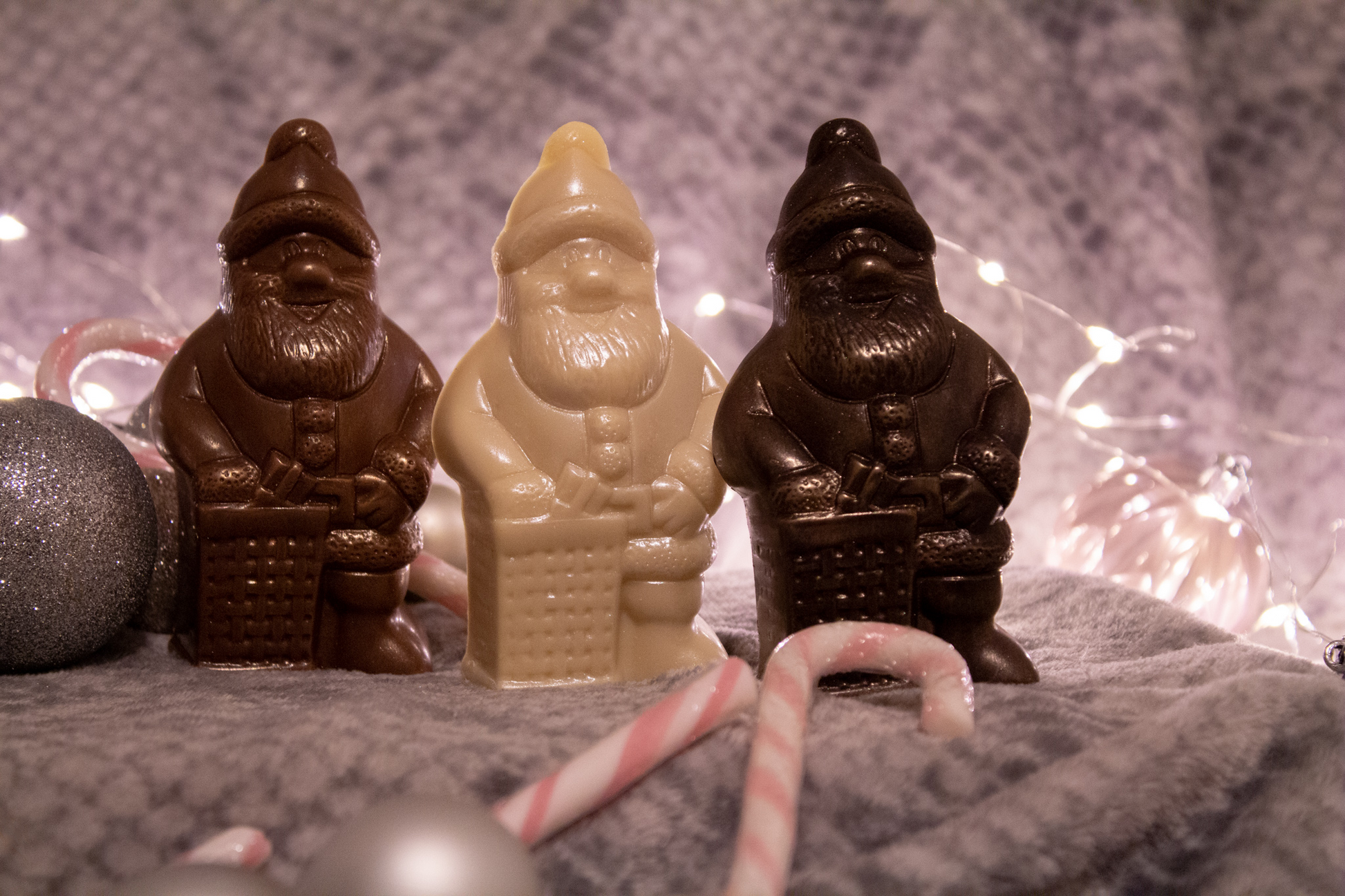 A set of three chocolate figurines in the shapes of Santa Claus, crafted in different colors representing milk, dark, and white chocolate, placed on a surface with holiday decorations around.