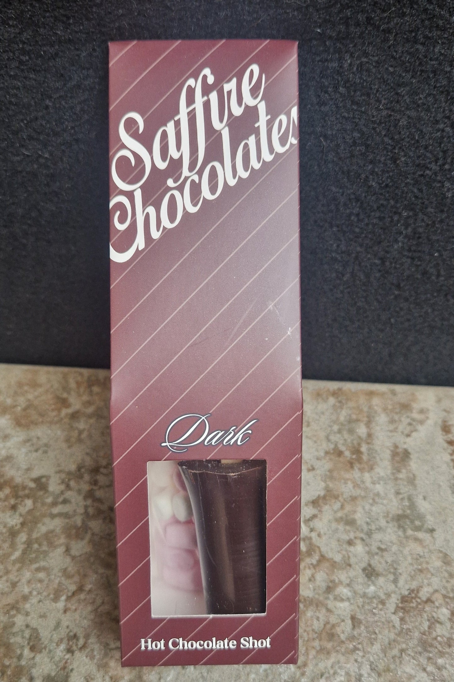 A boxed chocolate product labeled 'Saffire Chocolate' and 'Delic' with a small window showing a dark chocolate stick, placed on a split black and beige textured surface.
