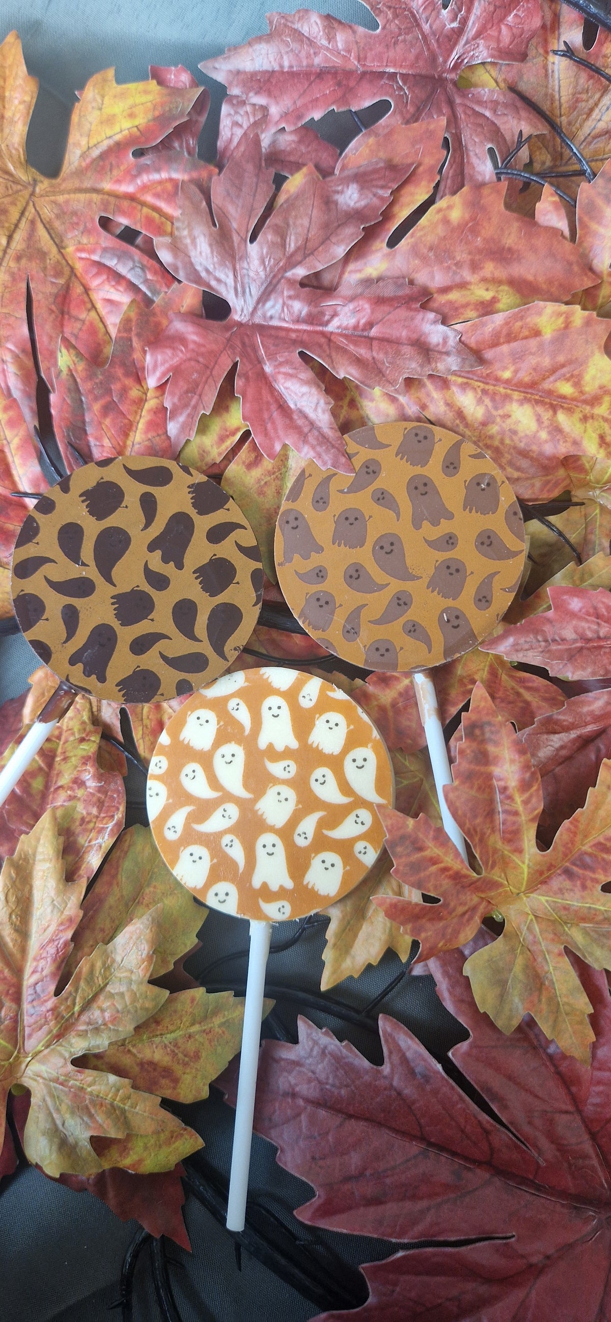 Three handmade Halloween chocolate lollies with ghost, bat and pumpkin designs on autumn leaves.