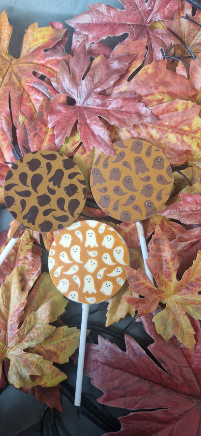 Three handmade Halloween chocolate lollies with ghost, bat and pumpkin designs on autumn leaves.