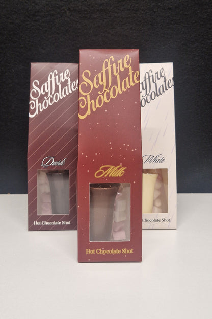 Three Saffire Chocolate Hot Chocolate Shot boxes standing upright on a white surface against a black background. The center box is dark red with gold lettering and shows a clear window revealing the chocolate shot inside.