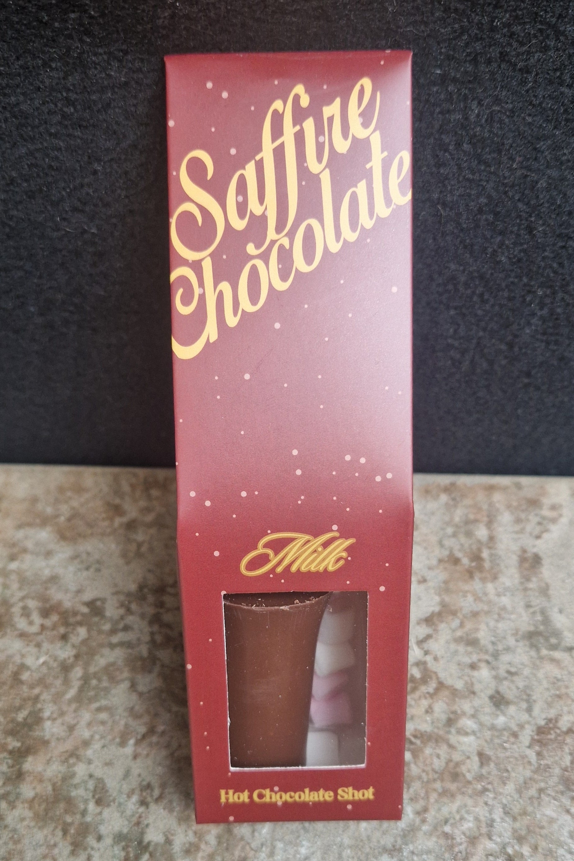 A boxed chocolate shot package standing upright on a textured beige surface against a dark background. The box is red with gold script reading 'Saffire Chocolate' and 'Milk Hot Chocolate Shot' near the bottom, with a clear window showing the chocolate shot inside.