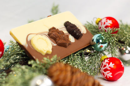 A chocolate bar with decorative elements resembling snow and holiday decorations, set against a background of Christmas decorations including red and green ornaments and brown branches.