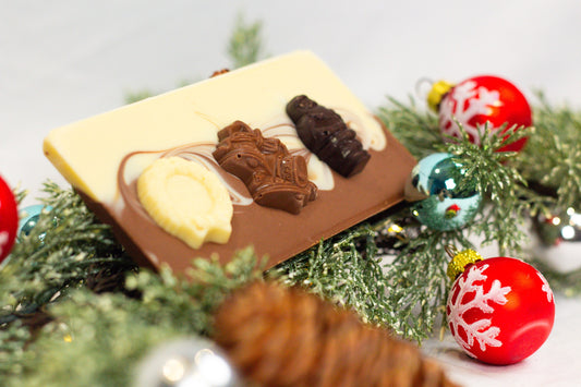 A chocolate bar with decorative elements resembling snow and holiday decorations, set against a background of Christmas decorations including red and green ornaments and brown branches.