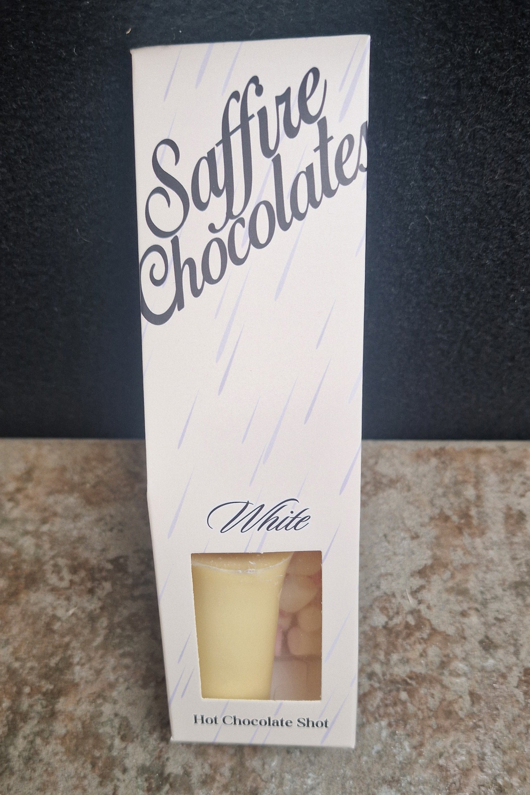 A tall white box labeled 'Saffire Chocolates' with a small clear window showing a pale yellow chocolate shot inside, standing on a speckled countertop against a dark background.