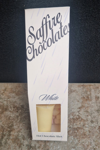 A tall white box labeled 'Saffire Chocolates' with a small clear window showing a pale yellow chocolate shot inside, standing on a speckled countertop against a dark background.