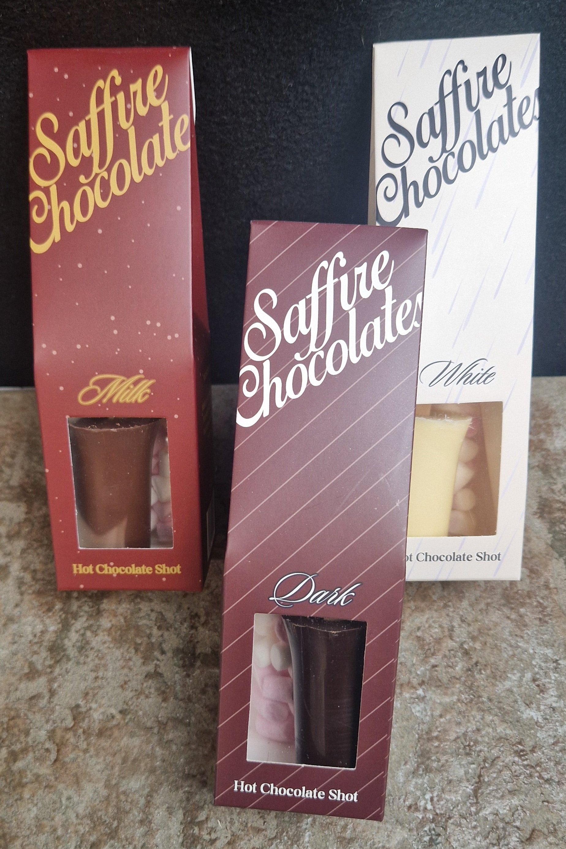 Three boxed chocolate shots labeled Saffire Chocolate in red, white, and dark brown packaging, standing on a speckled countertop against a dark background.