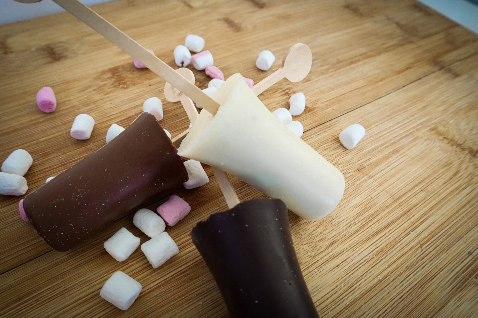 Three chocolate popsicles on a wooden surface with scattered marshmallows.