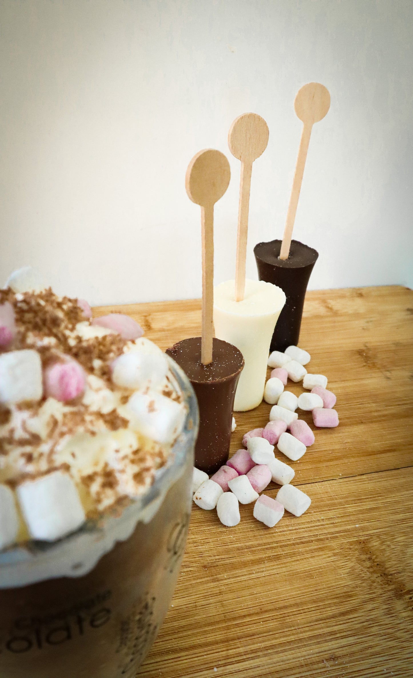 Hot chocolate with marshmallows, wooden stir sticks, and a hot chocolate shot on a wooden surface.