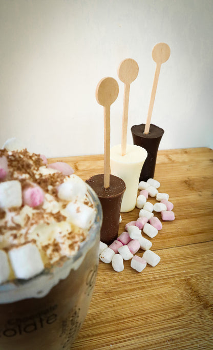 Hot chocolate with marshmallows, wooden stir sticks, and a hot chocolate shot on a wooden surface.